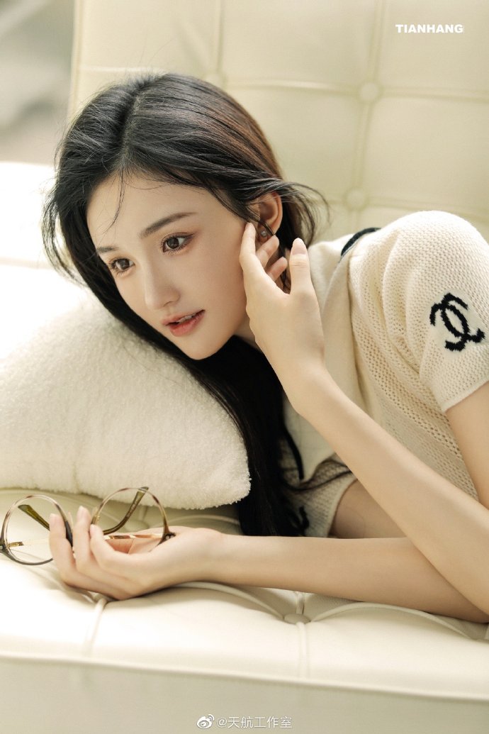 浅色短袖长发人物靠在白色靠枕边，手中拿着眼镜，后方为浅色软包面。